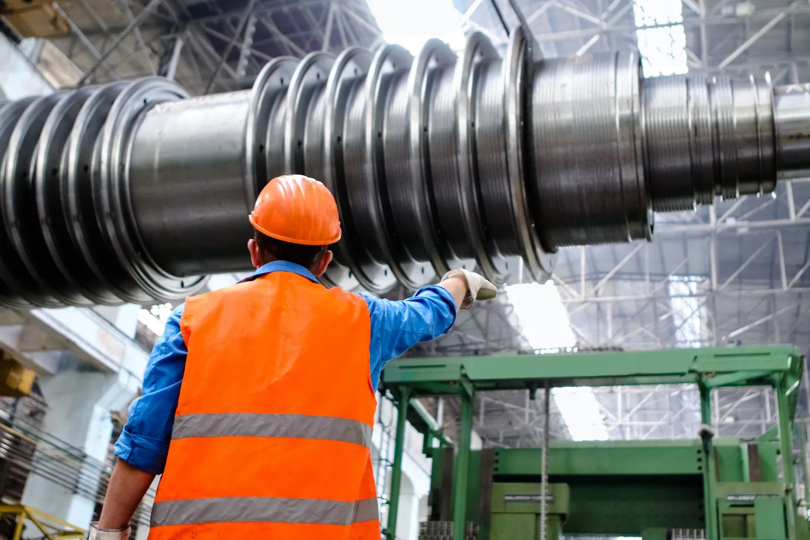 A technician in a hard hat and high-visibility vest inspecting a large machined rotor shaft in a factory hall