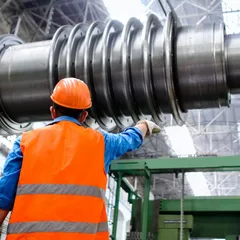 A technician in a hard hat and high-visibility vest inspecting a large machined rotor shaft in a factory hall