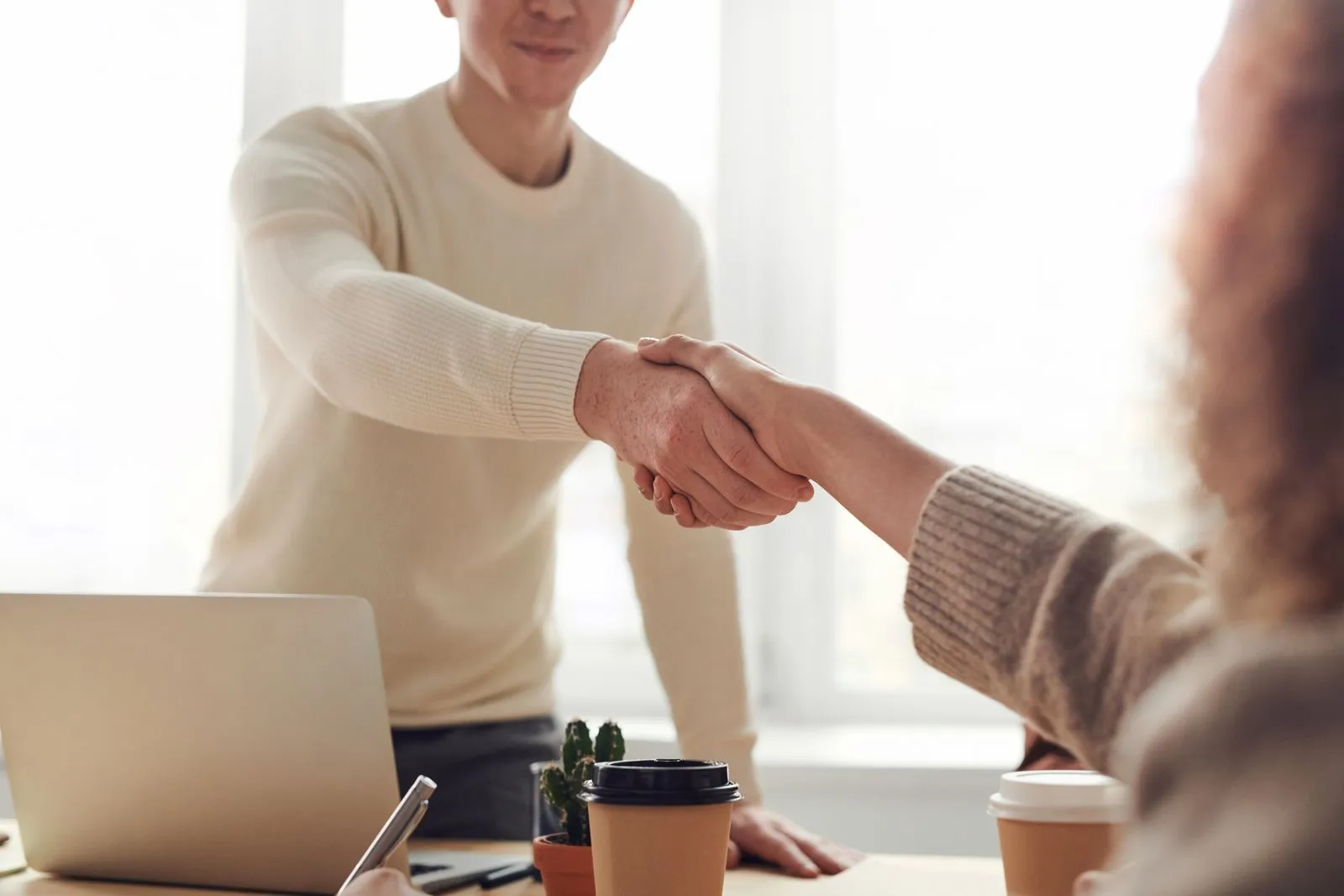Two people shaking hands across a desk in a bright office