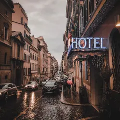 A rain-slicked cobbled street in Rome at dusk with a neon hotel sign