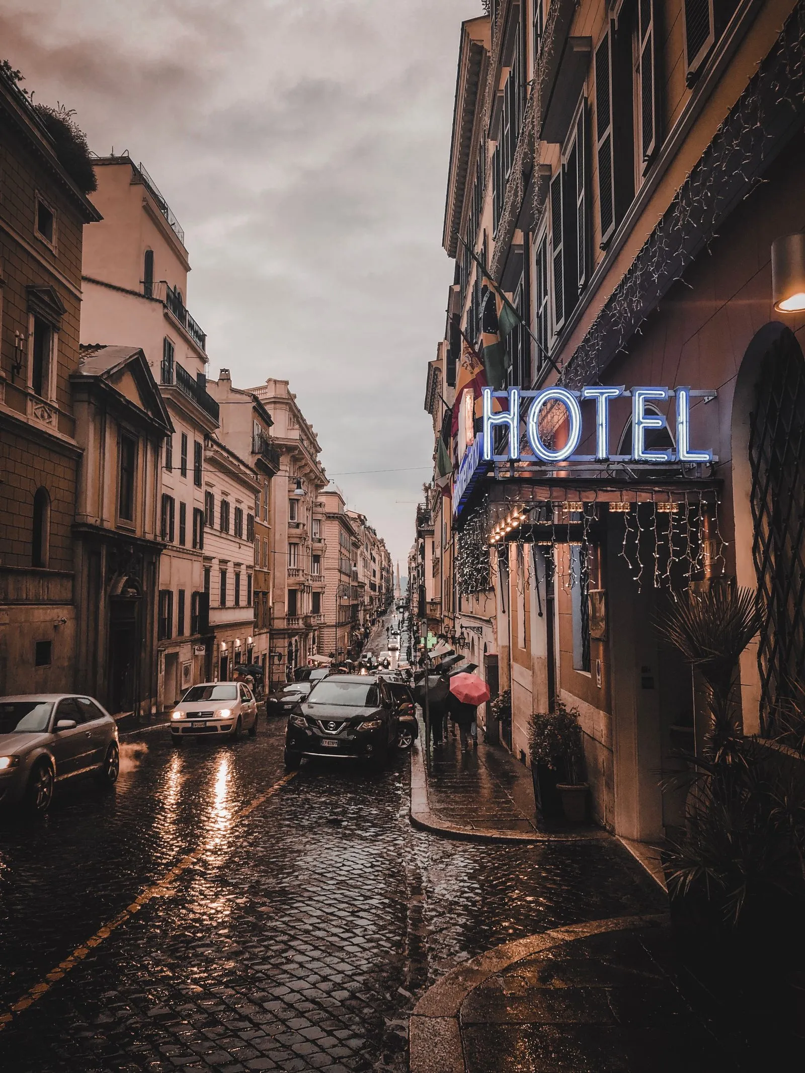 A rain-slicked cobbled street in Rome at dusk with a neon hotel sign