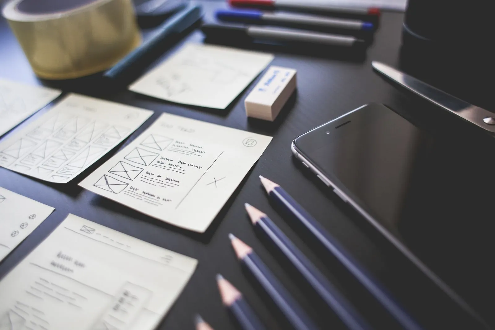 Hand-drawn interface wireframes, pencils and a phone arranged on a dark desk